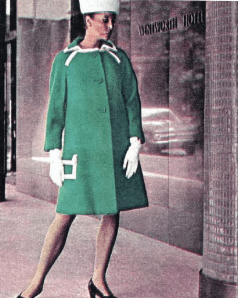 Woman in green coat, white hat, and heels walking down a sidewalk by a hotel building.