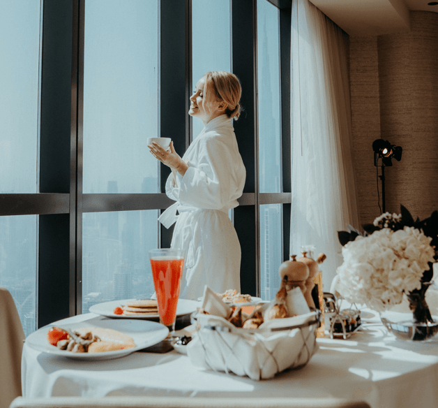 Woman in a white robe enjoys a peaceful breakfast by a large window with city views at Paramount Hotel Dubai
