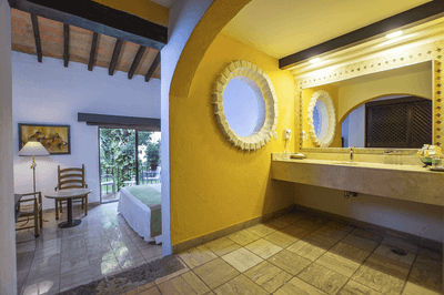 Yellow bathroom with double sink, circular mirror, and bed in adjacent room with sliding door and balcony.