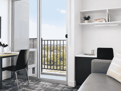 Modern student living room with desk, gray sofa, white shelving, and balcony access with city views at Student Living - Waymouth