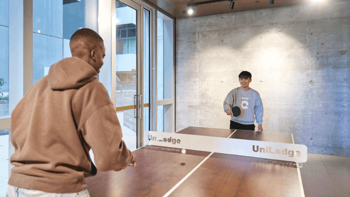 Two men play table tennis in a room with a UniLodge branded table, and a building in the background.
