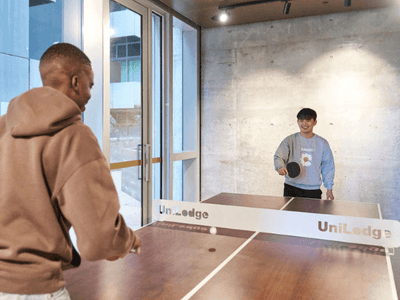 Two men play table tennis in a room with a UniLodge branded table, and a building in the background.