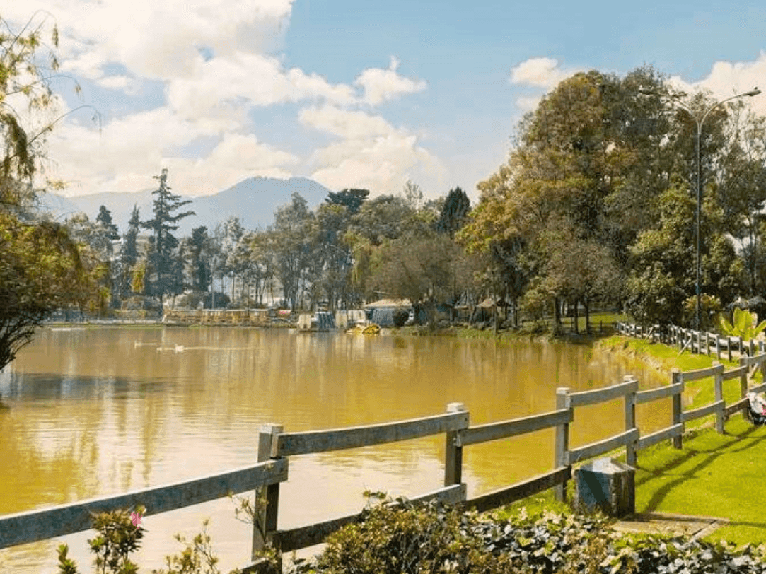 Vista panorámica de un lago sereno rodeado de un exuberante césped verde, árboles frondosos y una valla de madera, con montañas en el fondo bajo un cielo parcialmente nublado.