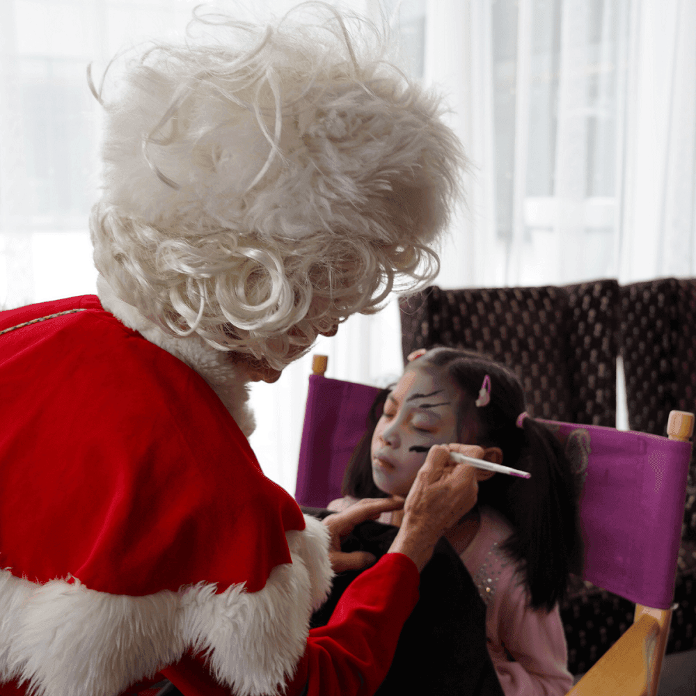 Santa painting a child\'s face as she sits in a purple chair by bright windows during festivities at Hotel X Toronto