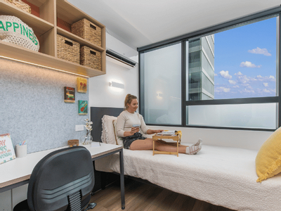 A woman sits on a bed in a room with a desk, chair, shelves, and a large window.
