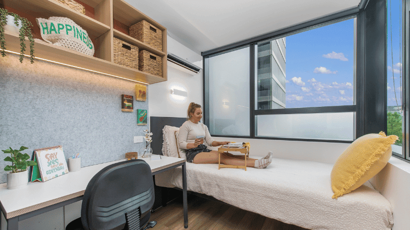 A woman sits on a bed in a room with a desk, chair, shelves, and a large window.