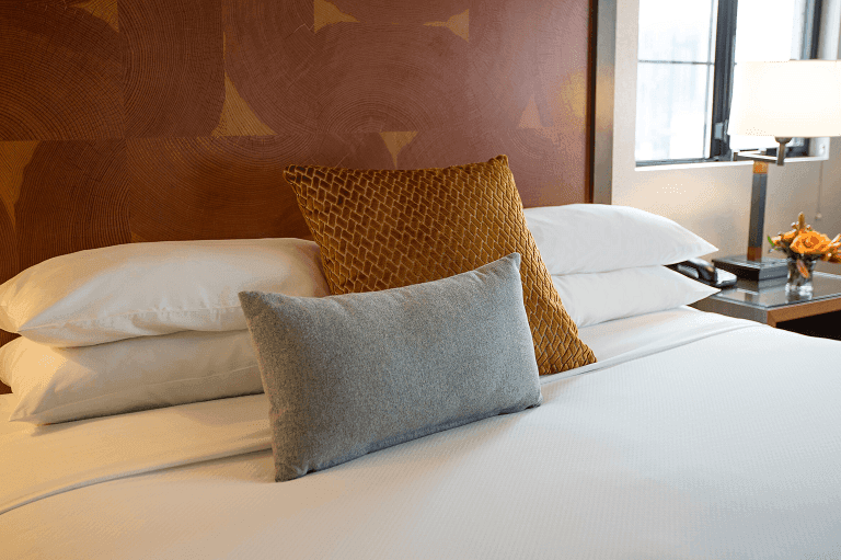 A neatly made bed with white sheets and several pillows, including a brown and a gray one, against a brown patterned wall, with a lamp and decorative items on a nightstand to the right.