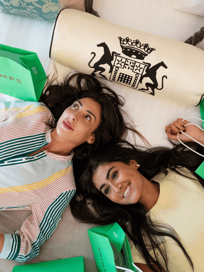 Two women lying on a bed with Printemps shopping bags, under a branded pillow at Hotel Westminster Paris