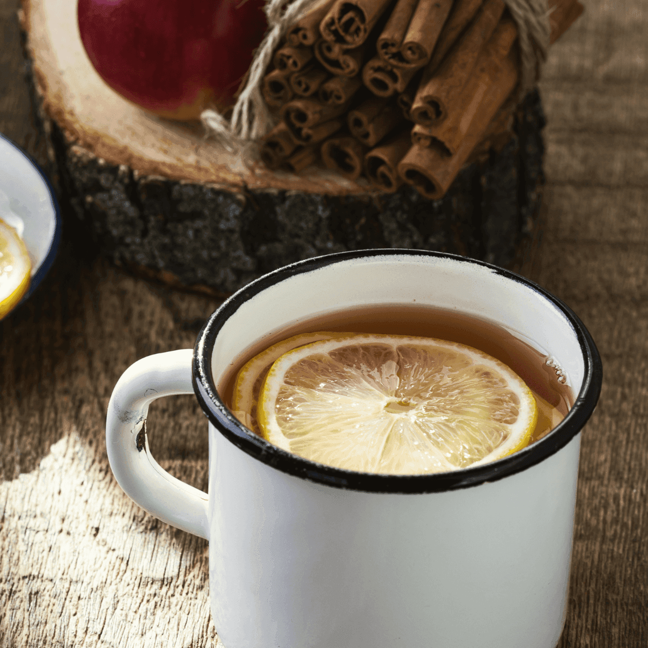 Mug of tea with lemon slices, cinnamon sticks, and apple slices on a wooden surface.