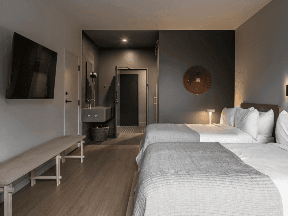 A room with two beds, a bench, a television, and a bathroom with a sink.