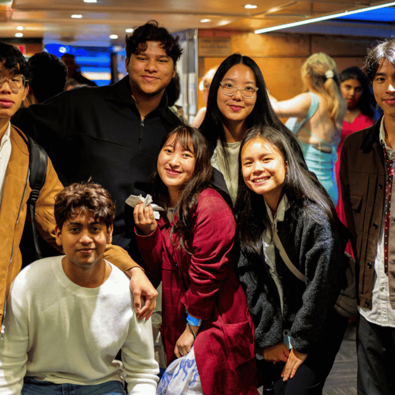 Group of seven young students smiling together in a lively indoor social space with UniLodge branding.