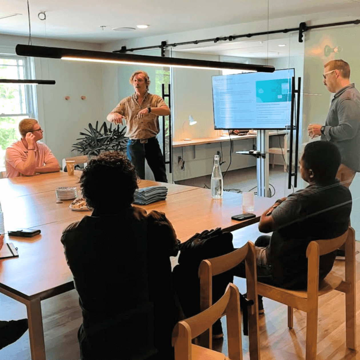 Group of people in a meeting room with two men standing and a large screen displaying content.