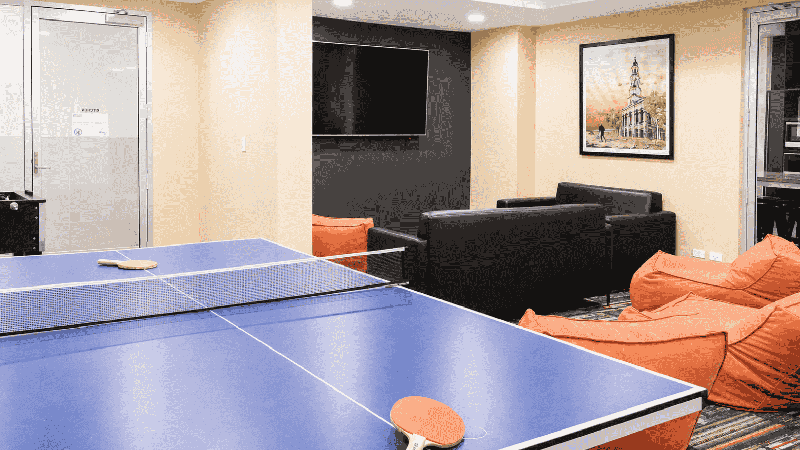 Modern recreation room with ping pong table, black leather sofa, orange bean bags, and wall-mounted television.