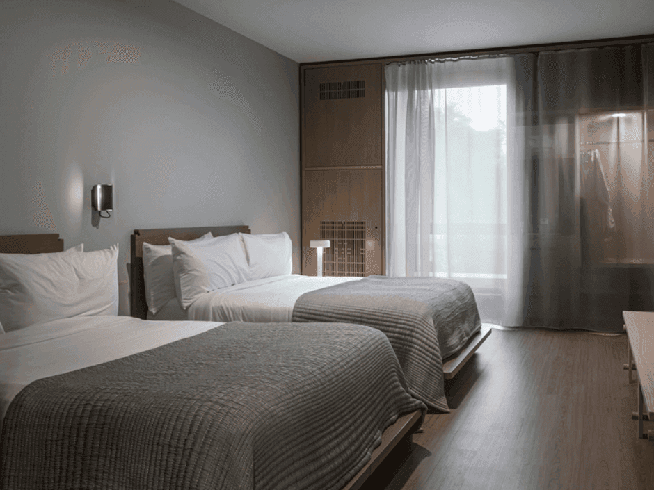 Two beds with white pillows and gray blankets, wooden floors, and a white curtain by the window.