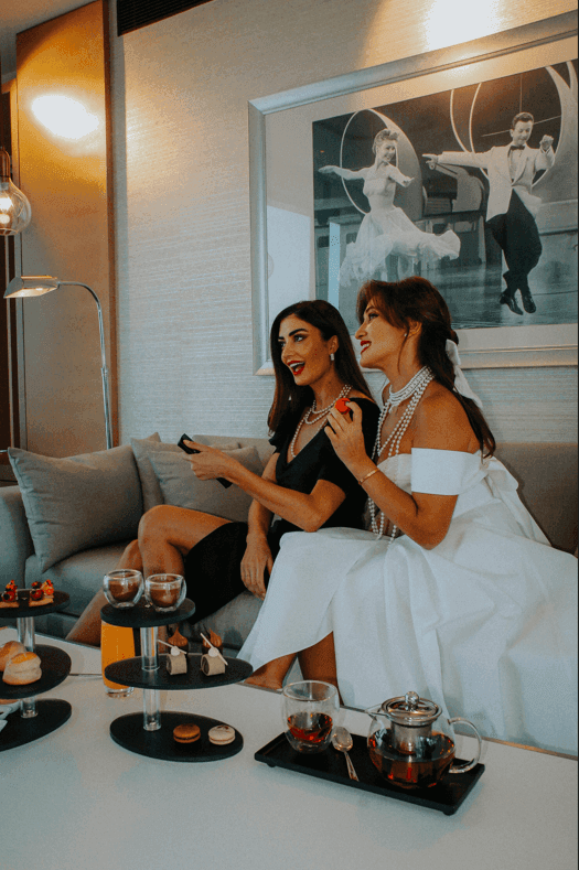 Two elegantly dressed women enjoying tea and treats while lounging on a plush sofa at the Paramount Hotel in Dubai