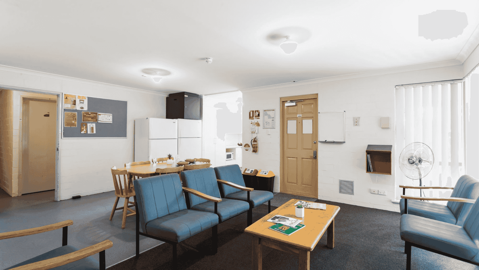 Comfortable lounge area with blue chairs, a wooden table, and a bulletin board at UniLodge at Curtin University - Vickery House.