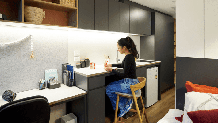 Woman sitting at a small kitchen counter writing with a pen in a compact living space.