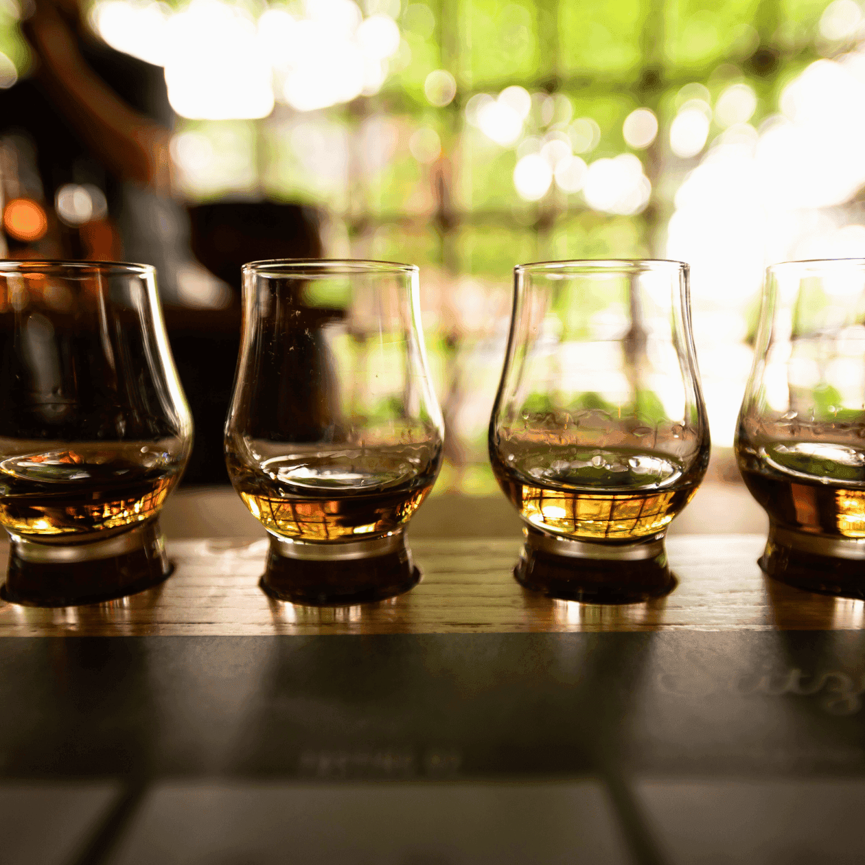 Four empty glasses lined up on a wooden table with a blurred background of green foliage and light.