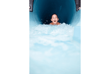 Boy smiling and splashing at the end of a blue water slide in the indoor pool facility at Rodd Hotels & Resorts
