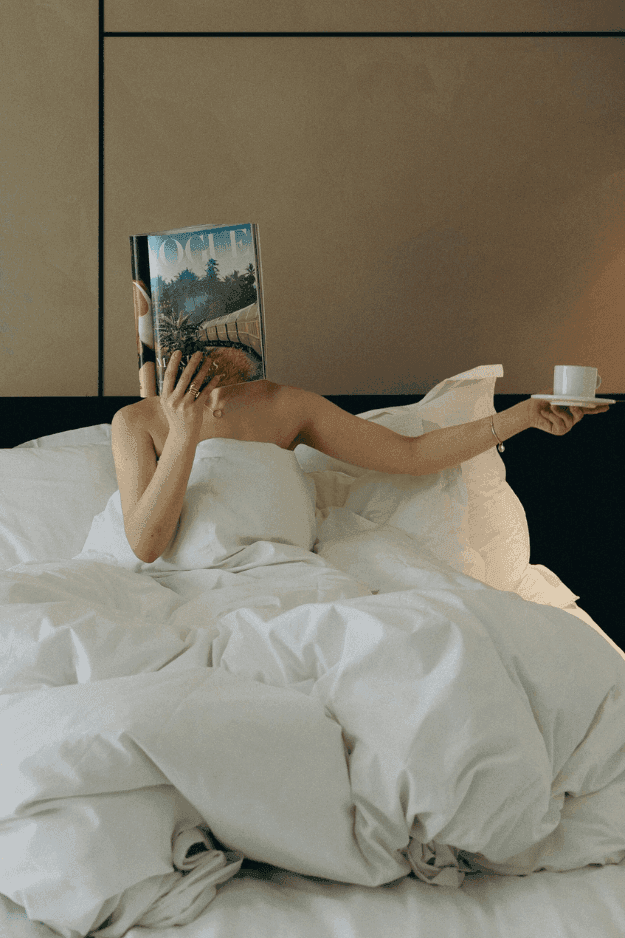 Woman relaxing in bed holding a coffee cup and a magazine, basking in morning light at The Rhyland Hotel in Flushing, NY