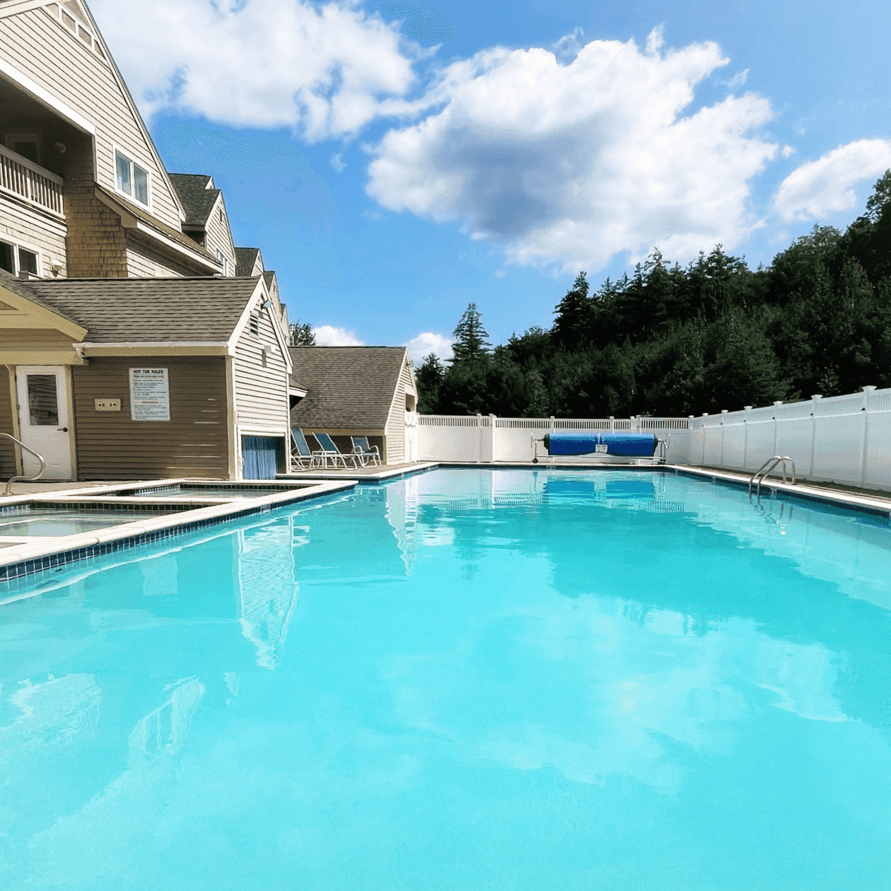Large heated pool with blue water and white fences, surrounded by trees and buildings under a partly cloudy sky.
