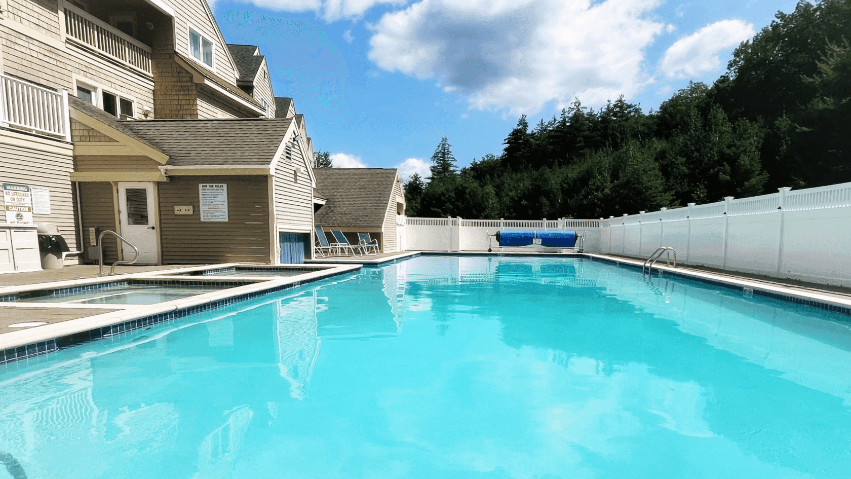 Large heated pool with blue water and white fences, surrounded by trees and buildings under a partly cloudy sky.