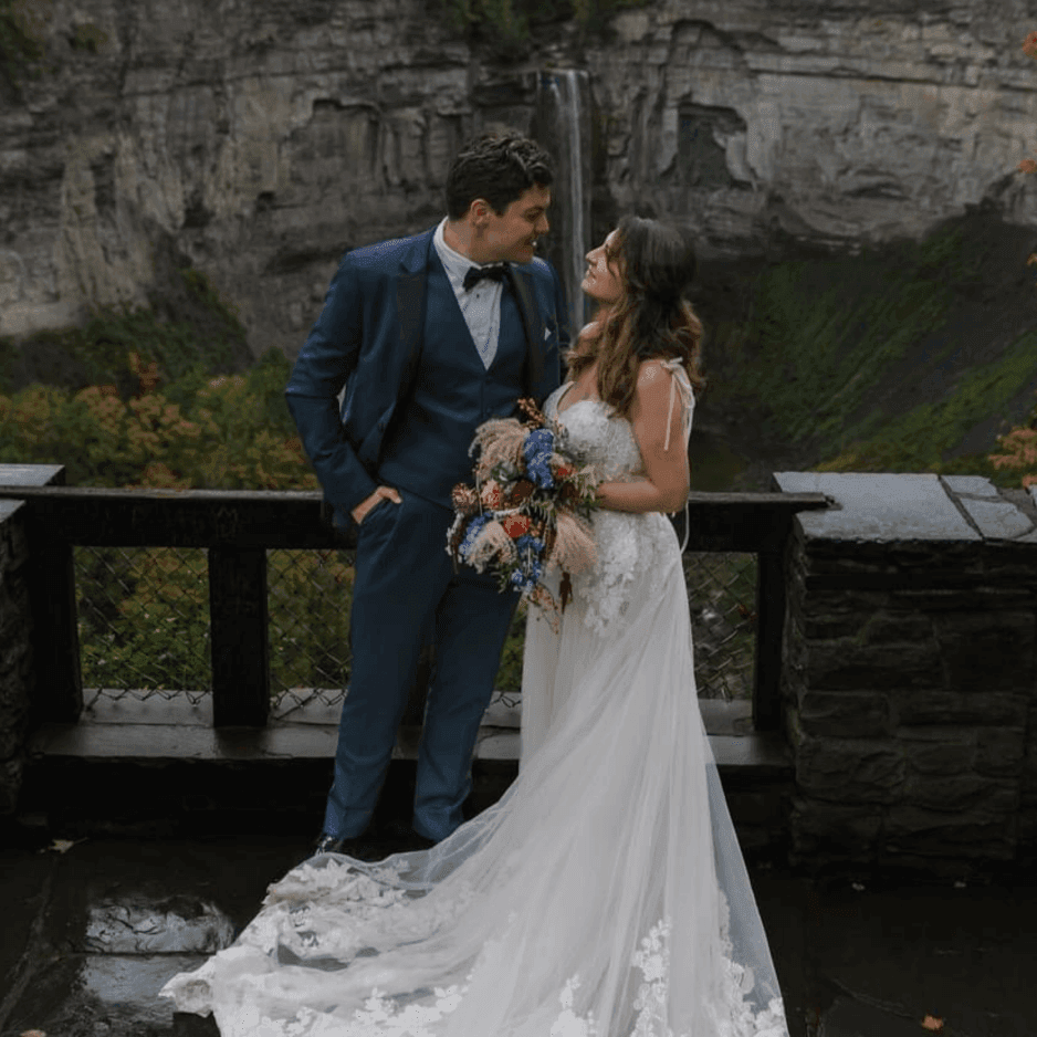 Bride and groom holding a colorful bouquet by a stone railing beneath a towering waterfall at La Tourelle Inn and Spa