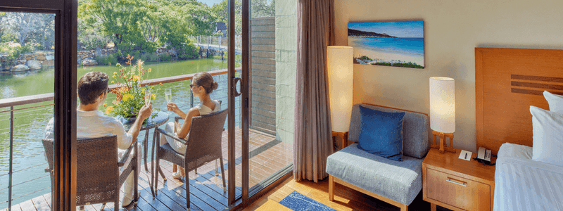 Couple enjoying a serene lakeside view from their private balcony with comfortable seating and a cozy bedroom nearby.