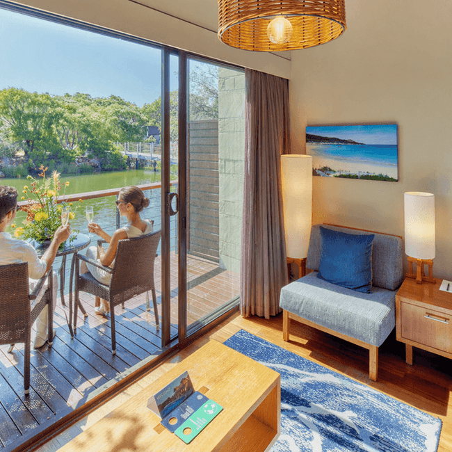 Couple enjoying a serene lakeside view from their private balcony with comfortable seating and a cozy bedroom nearby.