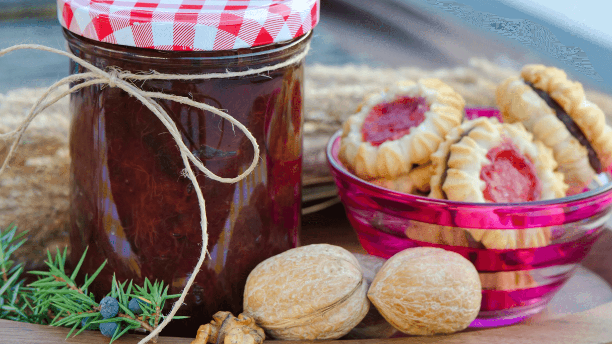 Homemade jam jar with checkered lid beside pink bowl of jam-filled cookies and walnuts at Busselton Foreshore Market.