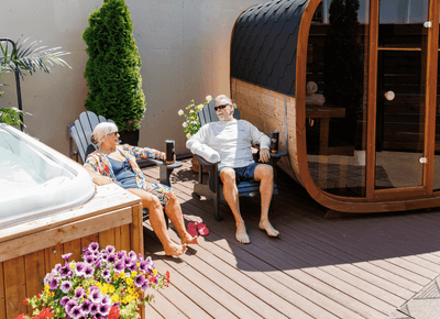 Couple resting in chairs between a bubbling hot tub and a barrel sauna on an outdoor sun patio deck at Rodd Moncton