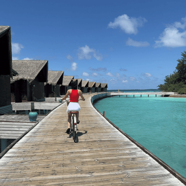 Woman in red top riding bicycle along wooden jetty between thatched water villas at Grand Park Kodhipparu Maldives