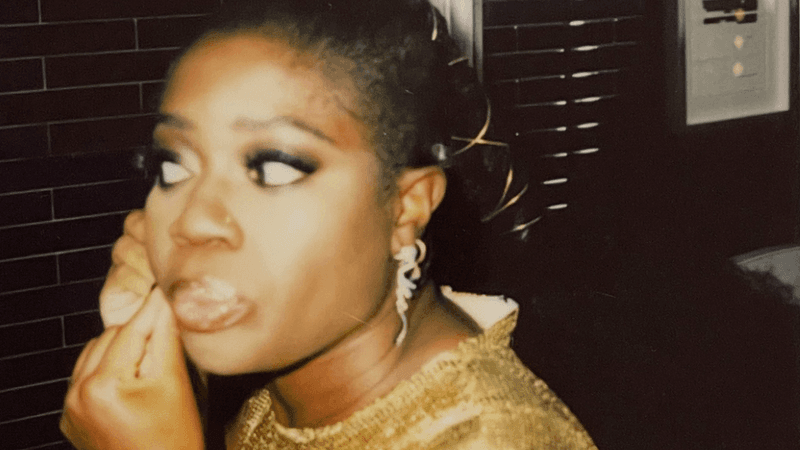 Clara Amfo wearing a gold dress and makeup, touching her face, in a Londoner bedroom.