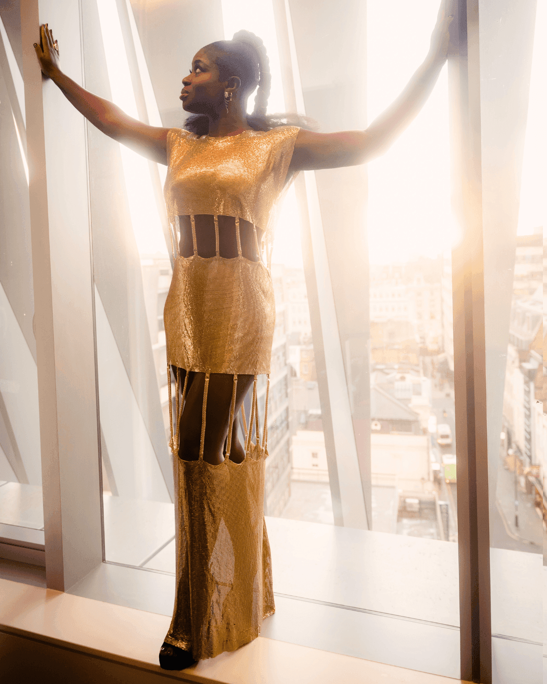 Stylish woman in a golden dress posing by a window with a city view at The Londoner