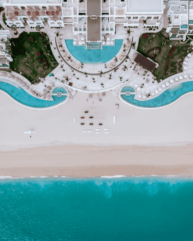 Aerial view of resort with curved pools, beach, palm trees, and white buildings.
