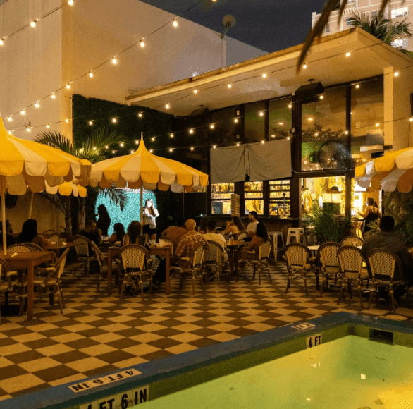 A nighttime view of an outdoor pool area with yellow umbrellas, string lights, and outdoor seating.
