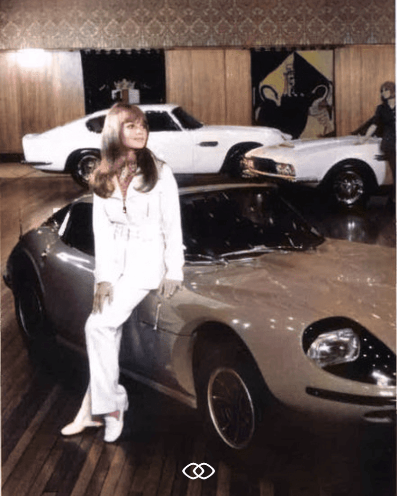 A woman in white sits on a silver sports car in a showroom with two other cars.