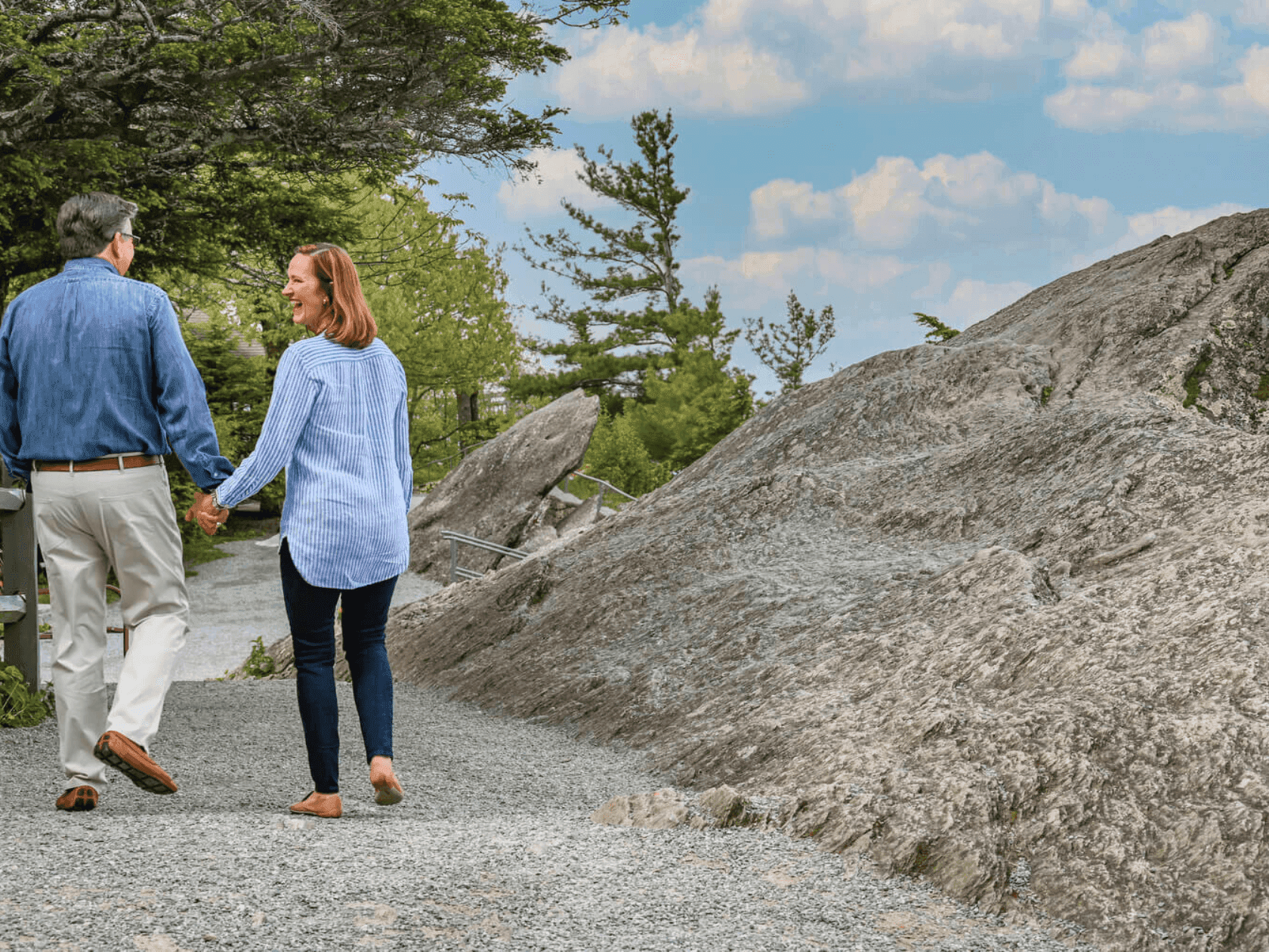 Elderly couple holding hands walking on a gravel path with rocky hill in the background.