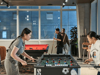 Two women play a foosball game while others observe in a room with large windows.