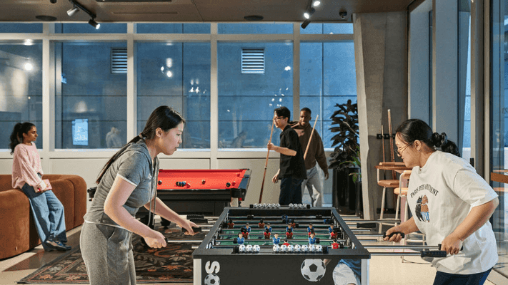 Two women play a foosball game while others observe in a room with large windows.