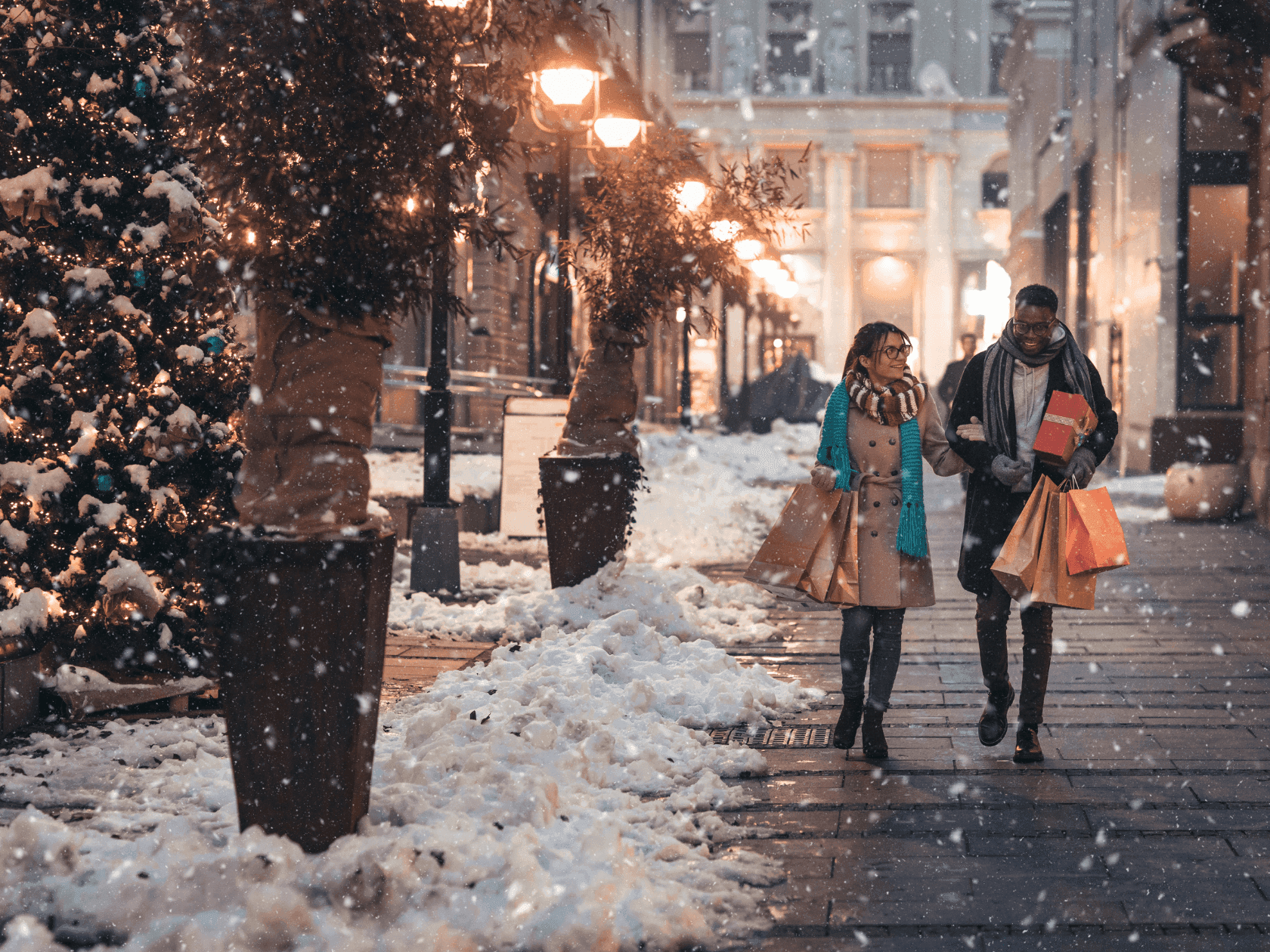 Couple walking in snow-covered city street with shopping bags, buildings, and lit streetlights on either side.