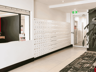 Modern reception area with white service counter, mailbox wall, tiled flooring, and potted plant at Student Living - Waymouth
