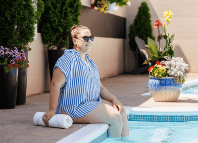 Woman wearing sunglasses sitting on the edge of an outdoor swimming pool beside vibrant potted flowers at Rodd Moncton