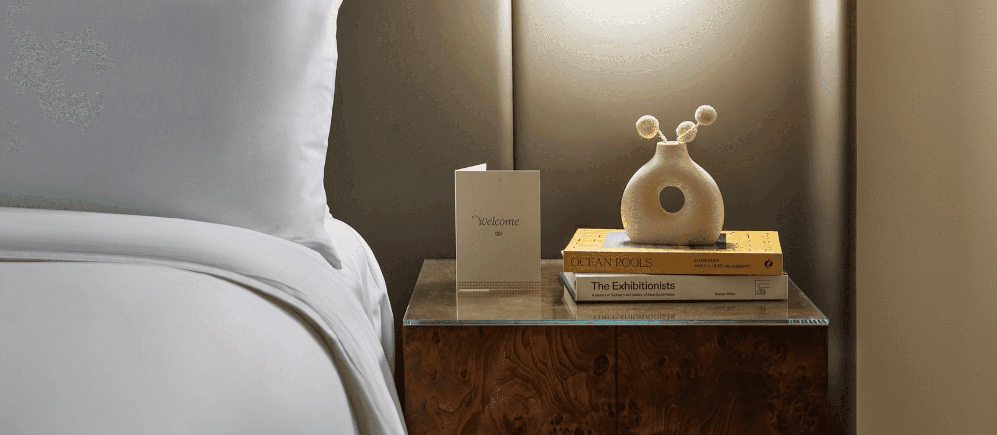 A hotel room with a bed, bedside table with books and vase, and a lit wall lamp.