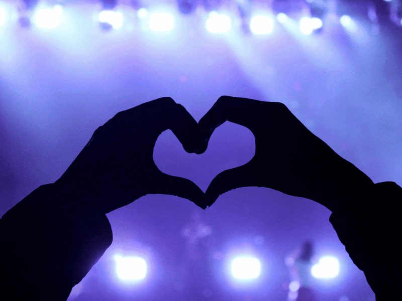 Silhouetted hands forming a heart shape against bright blue and purple concert stage lights.