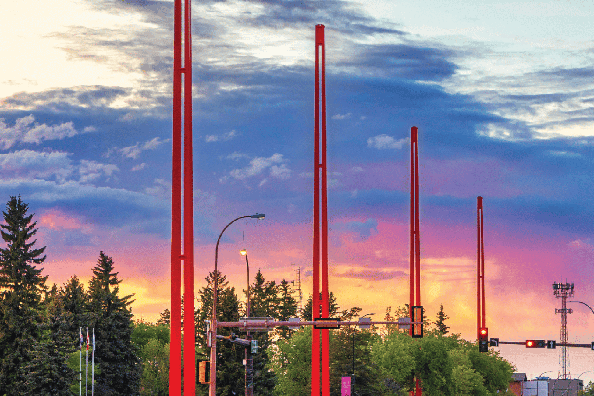 Photograph of tall red poles aligned along a street with traffic lights and trees under a colorful sunset sky. The scene highlights urban infrastructure elements against a vibrant backdrop of blue, purple, and orange hues.