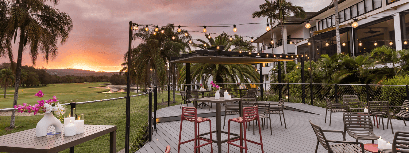 Outdoor dining area with tables, chairs, and string lights at sunset, overlooking a golf course and greenery.