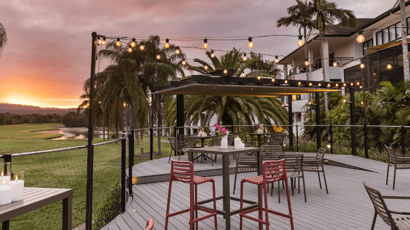 Outdoor dining area with tables, chairs, and string lights at sunset, overlooking a golf course and greenery.