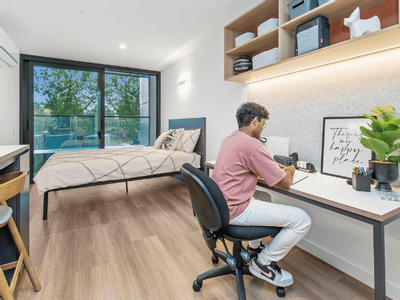 A man is seated at a desk in large apartment with a potted plant, and a bed is nearby.
