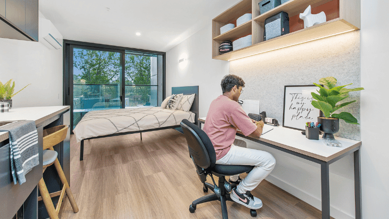 A man is seated at a desk in large apartment with a potted plant, and a bed is nearby.